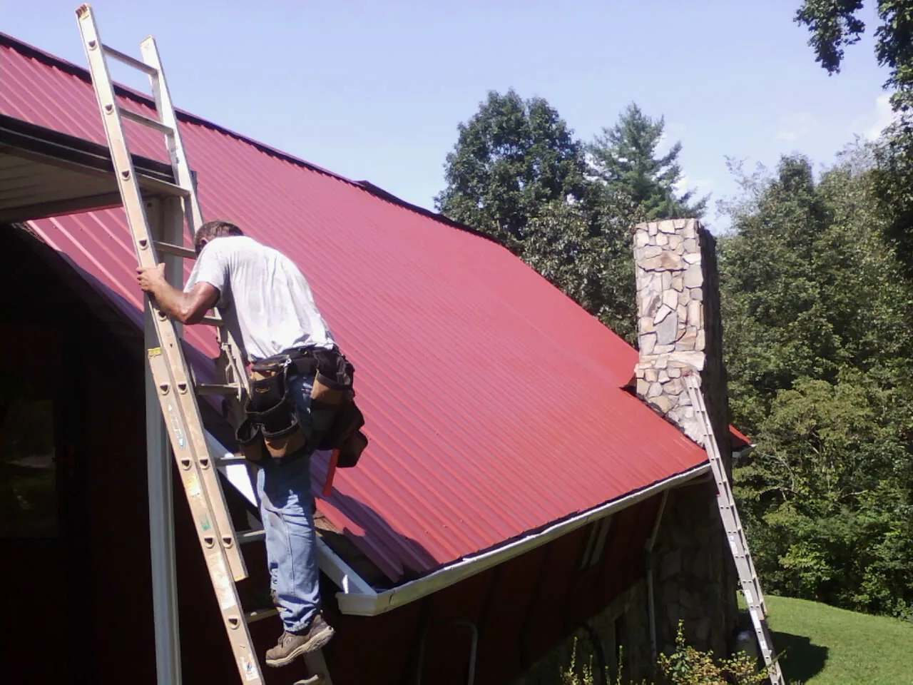Roofer on a ladder beside a red metal roof and stone chimney — Soffit & Fascia Repair by Roofing Brothers in Hamel